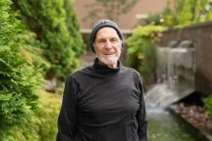 A man with a short grey beard and a black outfit smiles at the camera.