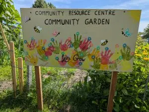 A hand-painted sign that reads "Community Resource Centre Community Garden." There are handprints created by the children along the bottom in green, pink, yellow and orange. There are some illustrations of bees and butterflies in the open spaces of the sign. it stands in front of a vegetable garden.