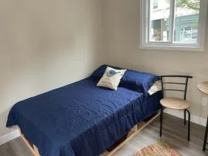 A bed sits against the wall of a tiny home. There is a window above the bed and it's covered in a dark blue coverlet. A small chair sits to the right of the bed.