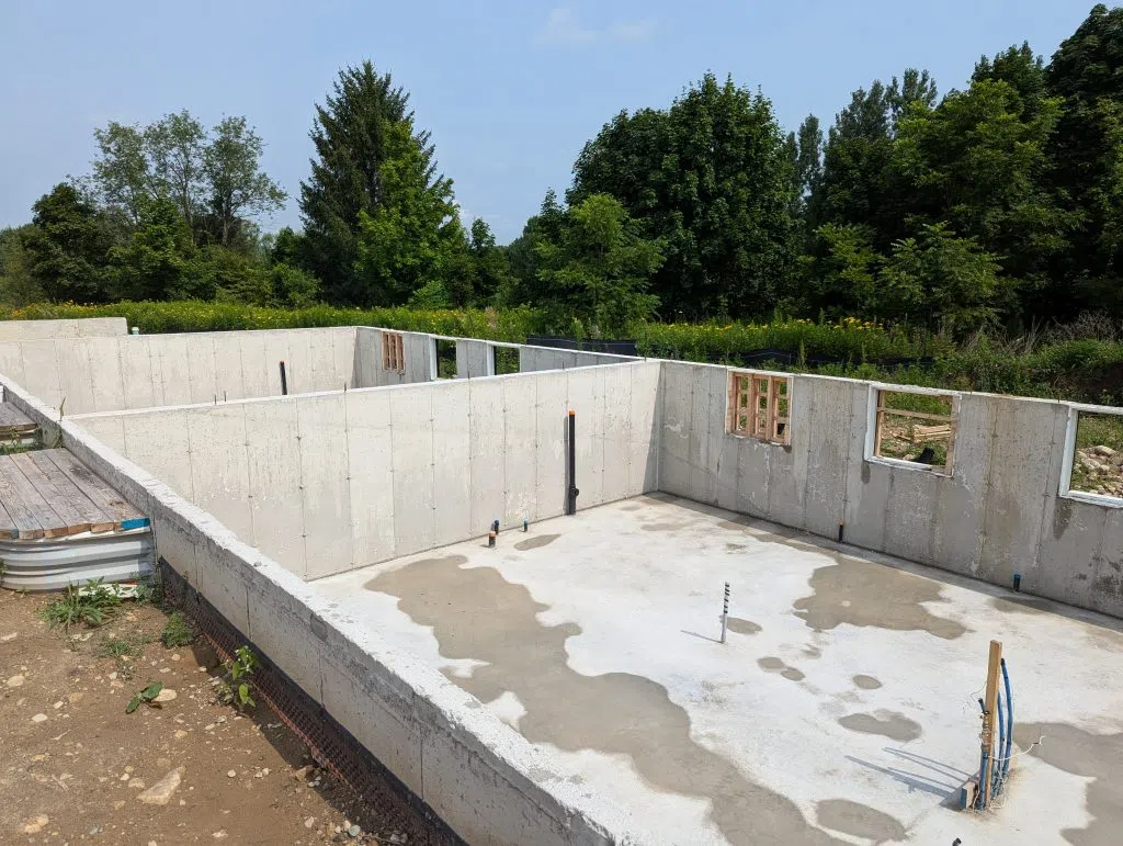 The foundation of two of the buildings is poured and electrical and plumbing rough-ins included. The image shows concrete walls and floor below grade. There is a stand of trees in the background. It is a sunny day.