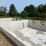The foundation of two of the buildings is poured and electrical and plumbing rough-ins included. The image shows concrete walls and floor below grade. There is a stand of trees in the background. It is a sunny day.
