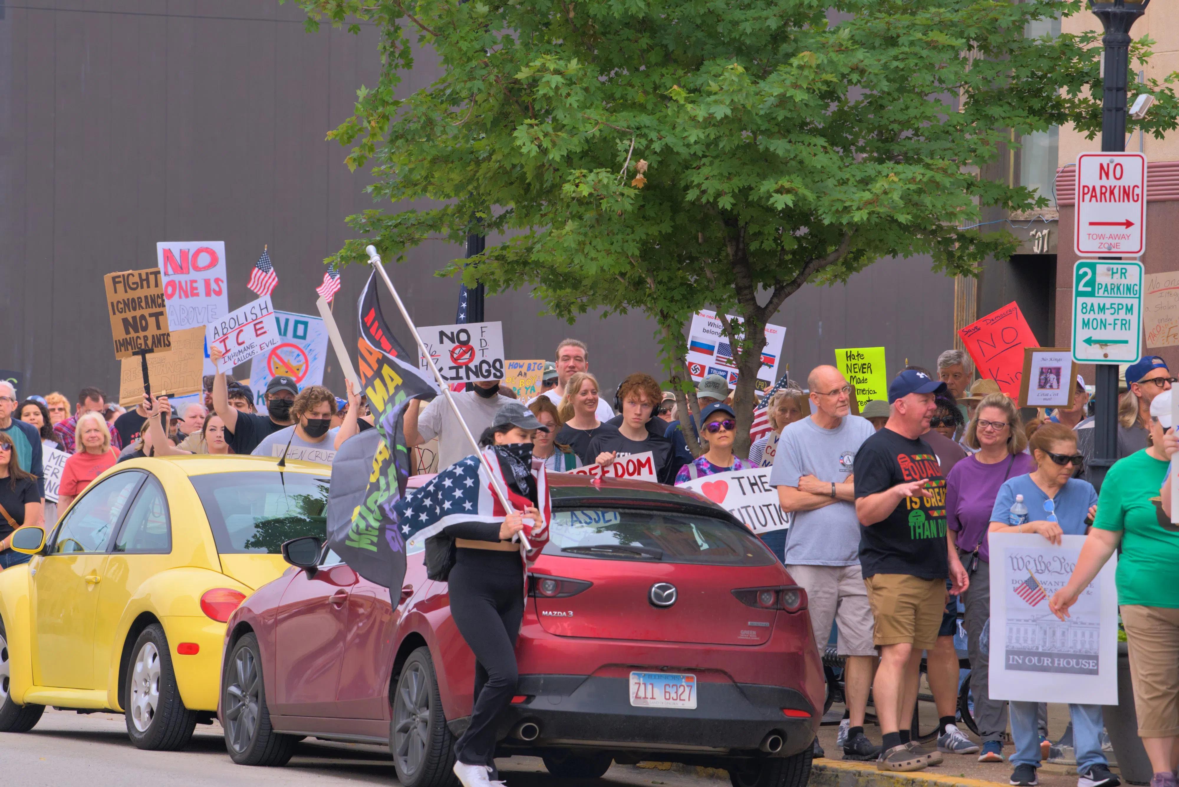 A protestor draped in US colors waves an anti-fascist pride flag as No Kings protesters wait to cross the street, June 14, 2025 (WMAY News, photo cred. Stan Anders)
