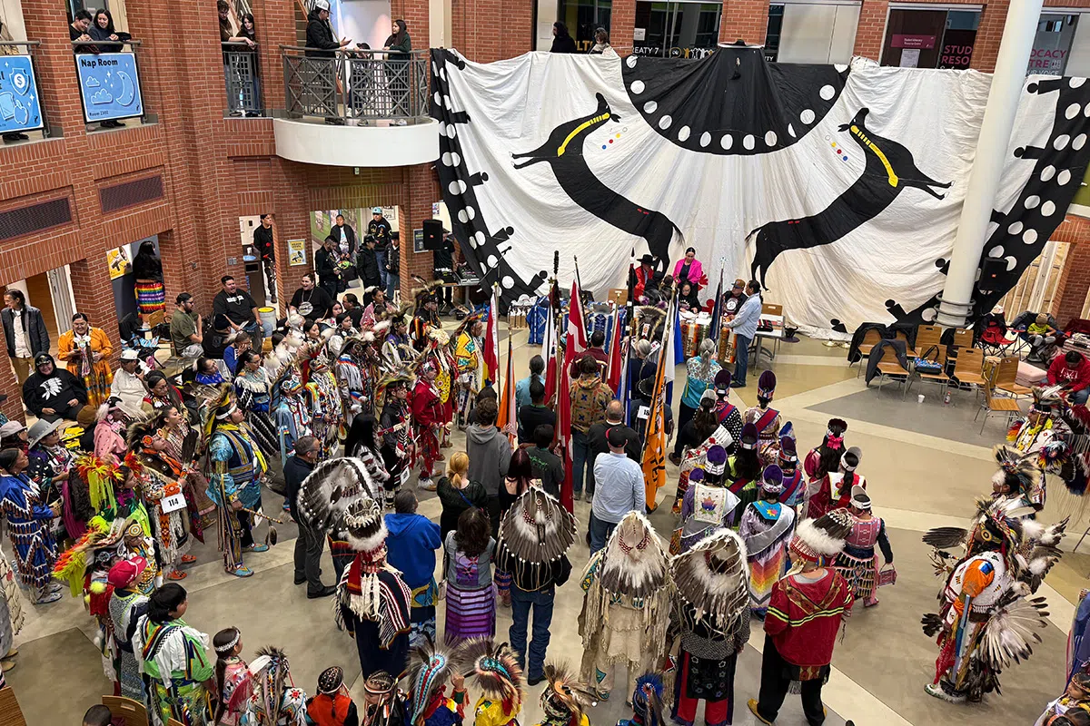 People gather indoors at MRU for a powwow, with dancers in colorful clothes standing in a large circle and a large banner hanging behind them.