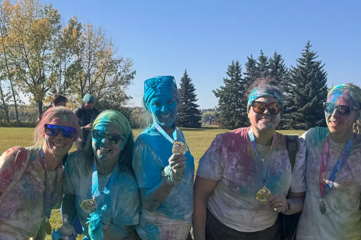 A group of women standing together on a sunny field after a colour run event, covered head-to-toe in colourful powder and wearing medals, smiling at the camera.