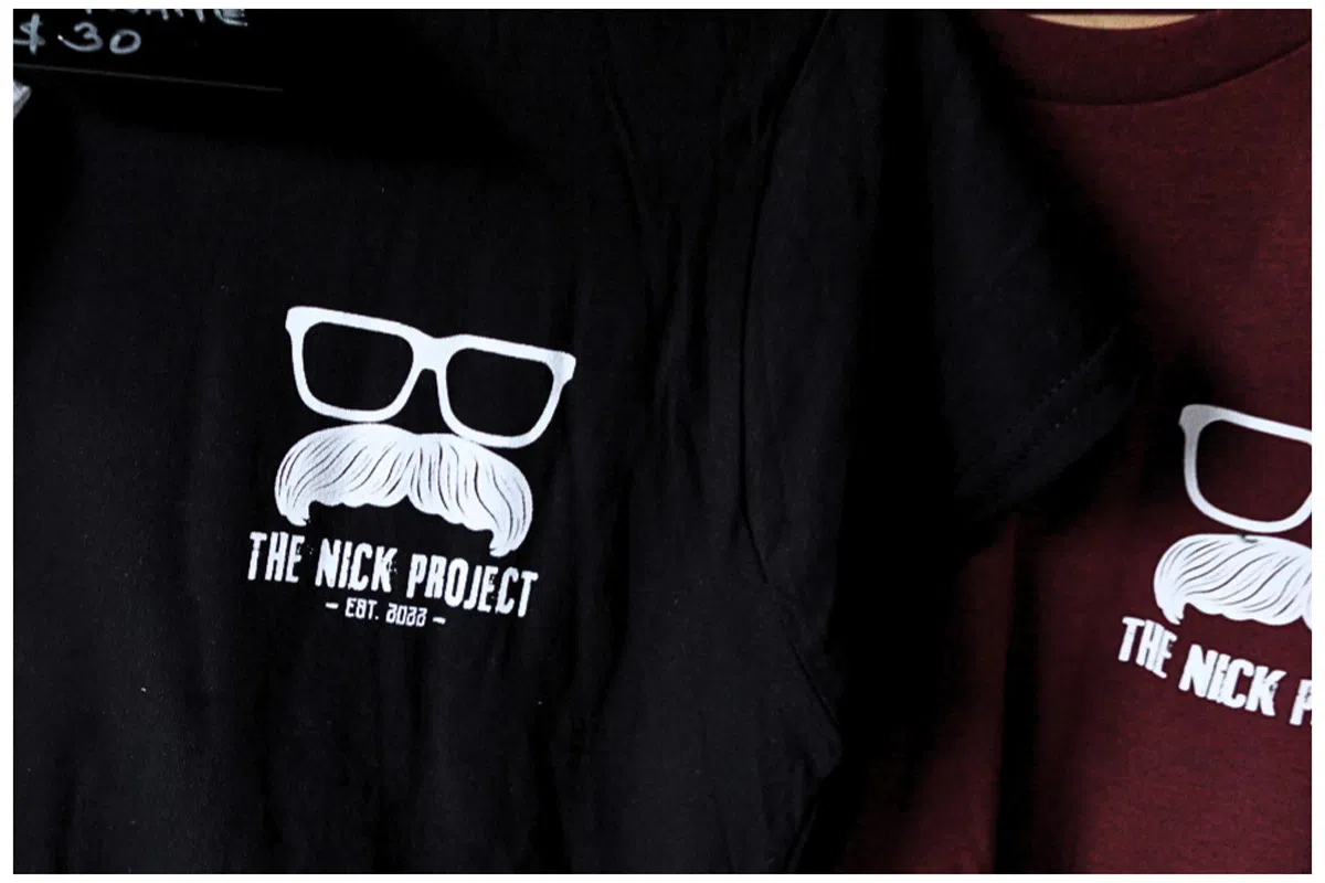 Black and maroon T-shirts hanging on display, each featuring a white graphic of glasses and a mustache with the text “The Nick Project – EST. 2022.”