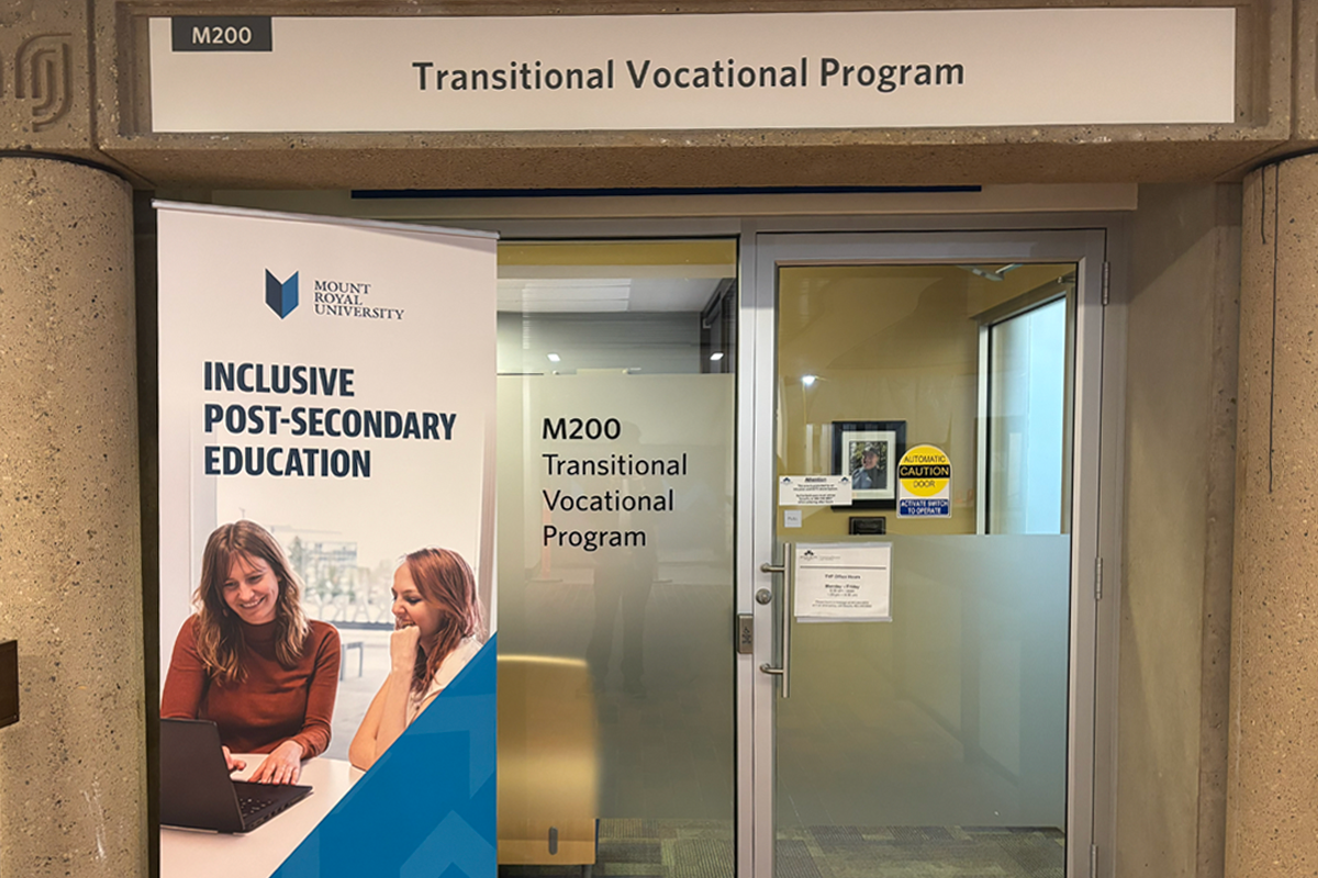 A banner reading Inclusive Post-Secondary Education with a picture of two women. The banner is next to a glass door reading M200, Transitional Vocational Program.