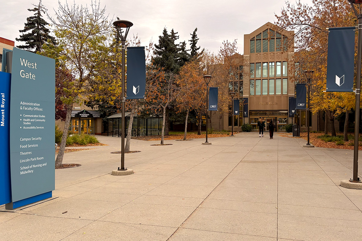 The exterior to the West Gate entrance of Mount Royal University. It is aligned with tree's on each side. The front of the building has a large window design on top. Photo is also accompanied with a sign on the right hand side stating West Gate listing key points of campus you can access through the West Gate entrance.