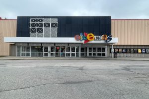 The front entrance of Canyon meadows theatre features a multi-coloured sign displaying the word "Cinemas", multiple glass doors for entrances and exits and movie posters displaying what movies are being featured at the cinema.