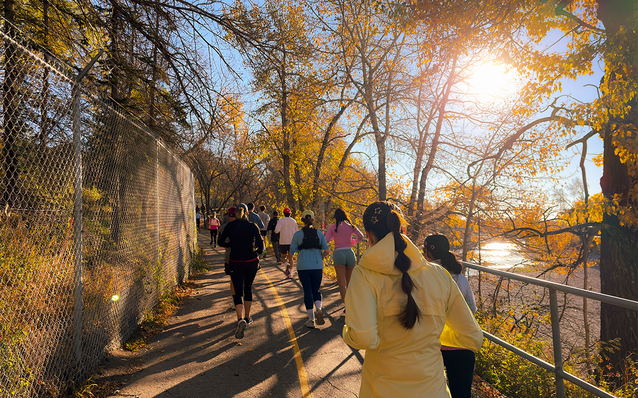 Runners participating in the First Responders Half Marathon race along the Bow River pathway in colourful outfits, running vests, and accessories like star-shaped hair clips. Orange fall leaves lines the path, with the river visible to their right and the Calgary zoo to their left.