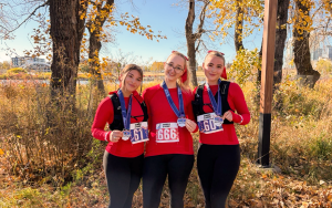 Three women in matching red long sleeve tops, running vests, metals and bedazzled race bibs stand at the end of the half marathon course, smiling and holding up their metals to the camera, surrounded by leaves and trees. with the Saint Patricks Park in the background.