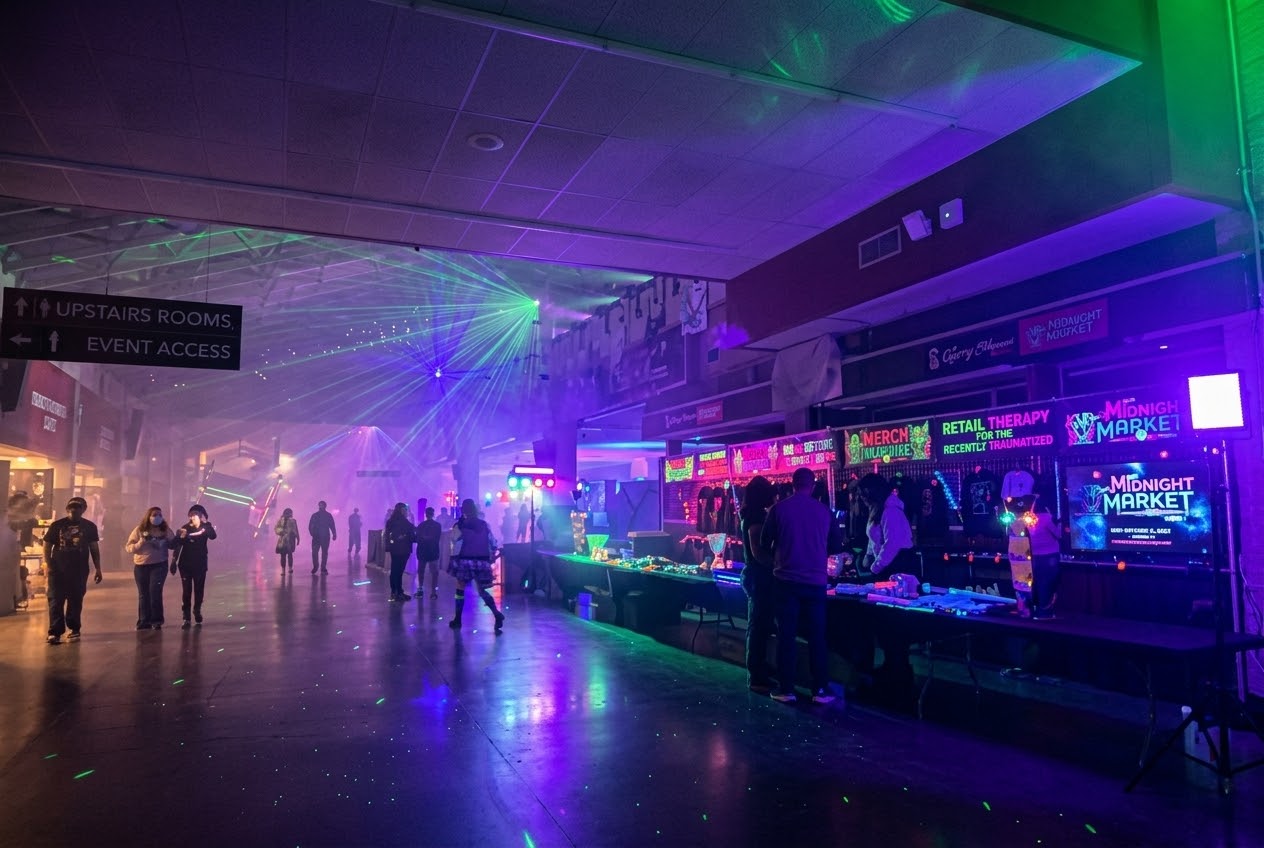 A dimly lit indoor hall filled with green lasers and purple neon lighting, showing visitors browsing merchandise tables labeled Midnight Market and signs reading Retail Therapy for the Recently Traumatized.