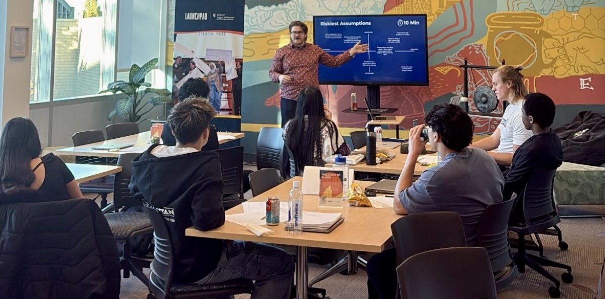 A man in a patterned shirt stands at the front of a university classroom with a large, colourful mural on the wall, gesturing toward a TV screen displaying a presentation slide titled 'Riskiest Assumptions.' Several students are seated at tables, facing the presentation.