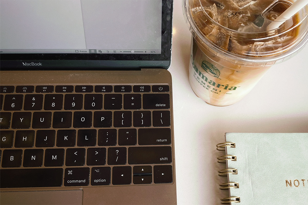 A close up horizontal image featuring a laptop, notebook, and an iced latte from Qamaria Yemen Coffee.