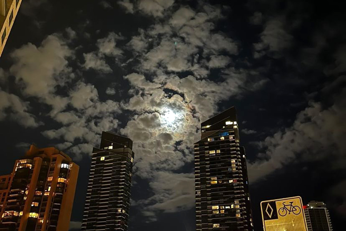 An exterior picture of the moon peeking out from behind the clouds and tall buildings, taken at night.
