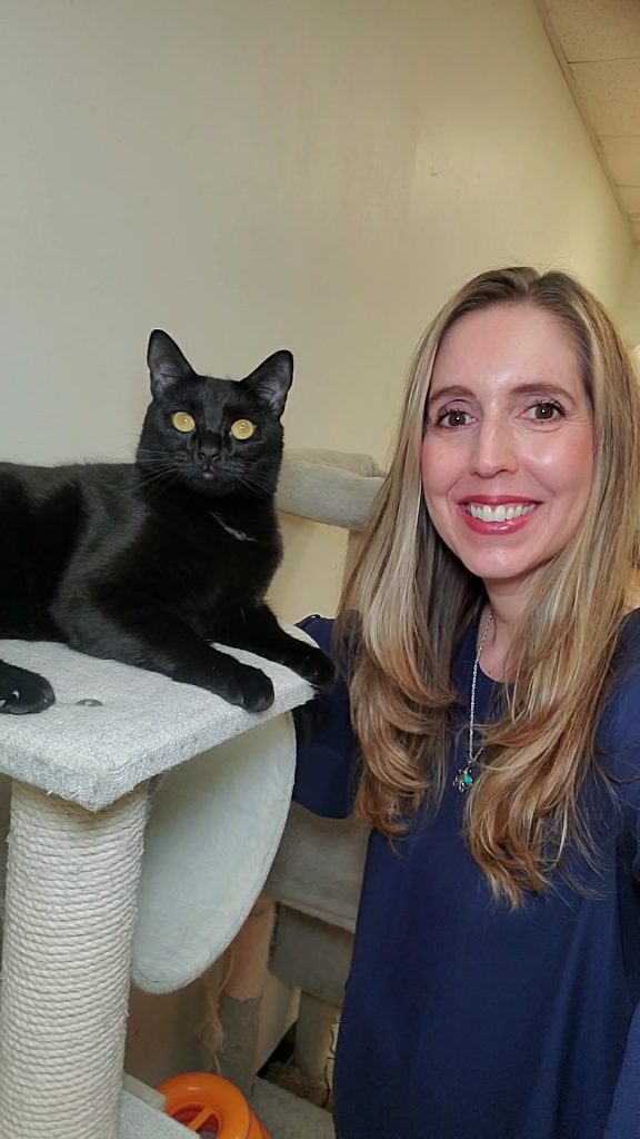 A blonde woman smiles next to a black cat with golden eyes