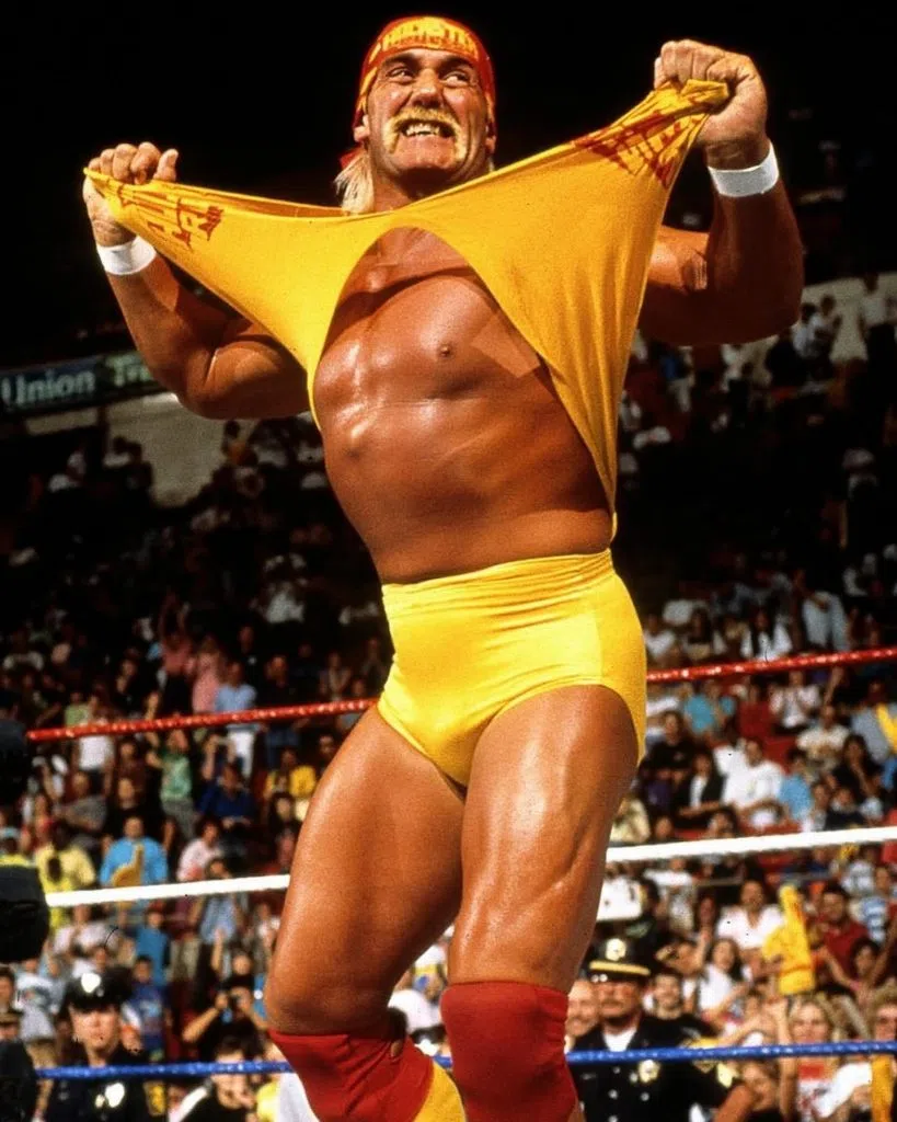 Hulk Hogan in yellow wrestling gear rips his shirt open in front of a cheering crowd during a WWF match.