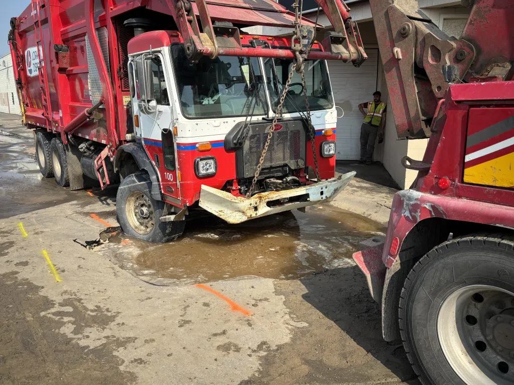 Tow truck preparing to pull a garbage truck out of a large sinkhole while workers stand nearby.