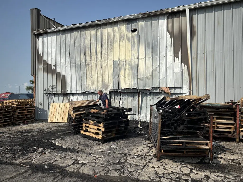 Man examines burnt wooden pallets stacked on a scorched asphalt lot outside a metal building wall showing smoke stains and fire damage at Auto Parts Plus in Regina.