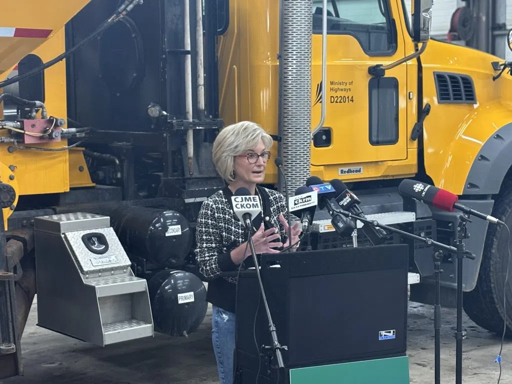 Shantel Lipp speaking at a podium with microphones in front of a yellow Ministry of Highways truck during the Orange Zone Construction Update announcement.