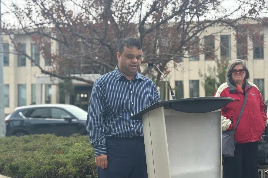 A man in a striped shirt speaks from a podium outdoors with people listening behind him during Regina’s PedApp unveiling.