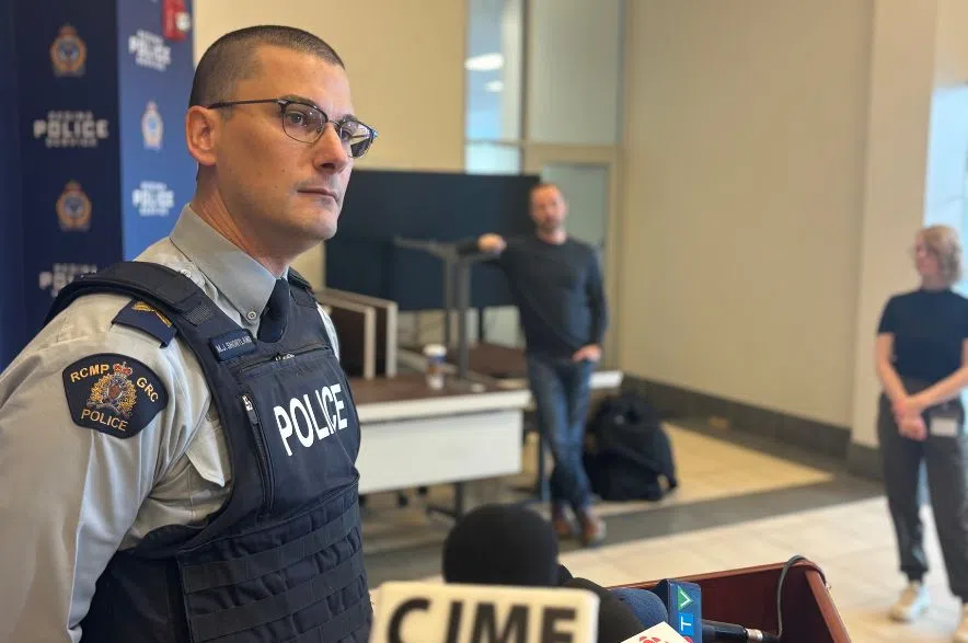 A uniformed Staff Sgt Michael Shortland with the RCMP stands at a podium during a media briefing on recent “grandparent scam” fraud cases. Microphones from CJME, CKOM and other outlets are visible in the foreground.
