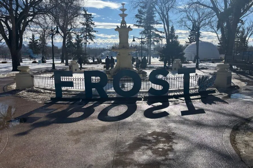 Large “FROST” letters in front of a fountain at a winter festival, with melting snow and puddles visible on the ground under a sunny blue sky.
