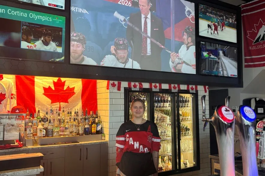 Addison White stands behind the bar at Boston Pizza in Regina beneath multiple TV screens showing Canada’s men’s hockey team at the Milano Cortina 2026 Winter Olympics.