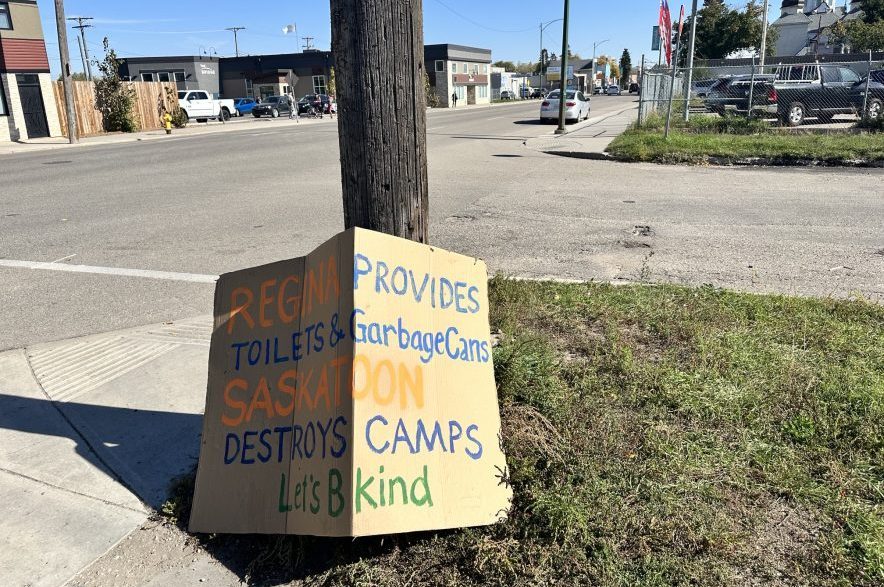 A piece of cardboard near the Saskatoon encampment on 20th Street at Avenue K reads "Regina provides toilets & garbage cans. Saskatoon destroys camps. Let's B kind."