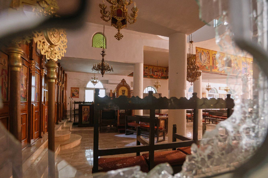 The interior of a Greek Orthodox church is seen through a shattered window.