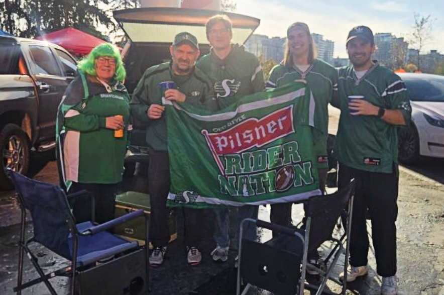 Five Saskatchewan Roughriders fans wearing green-and-white gear pose with a Rider Nation flag.
