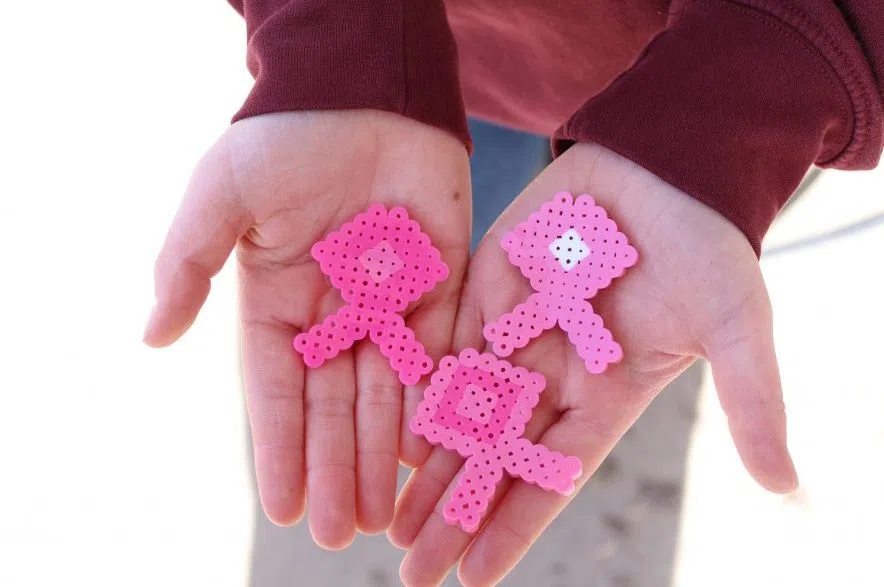 Lexi Curren holds three pink breast cancer awareness ribbons made of Perler beads in her outstretched hands
