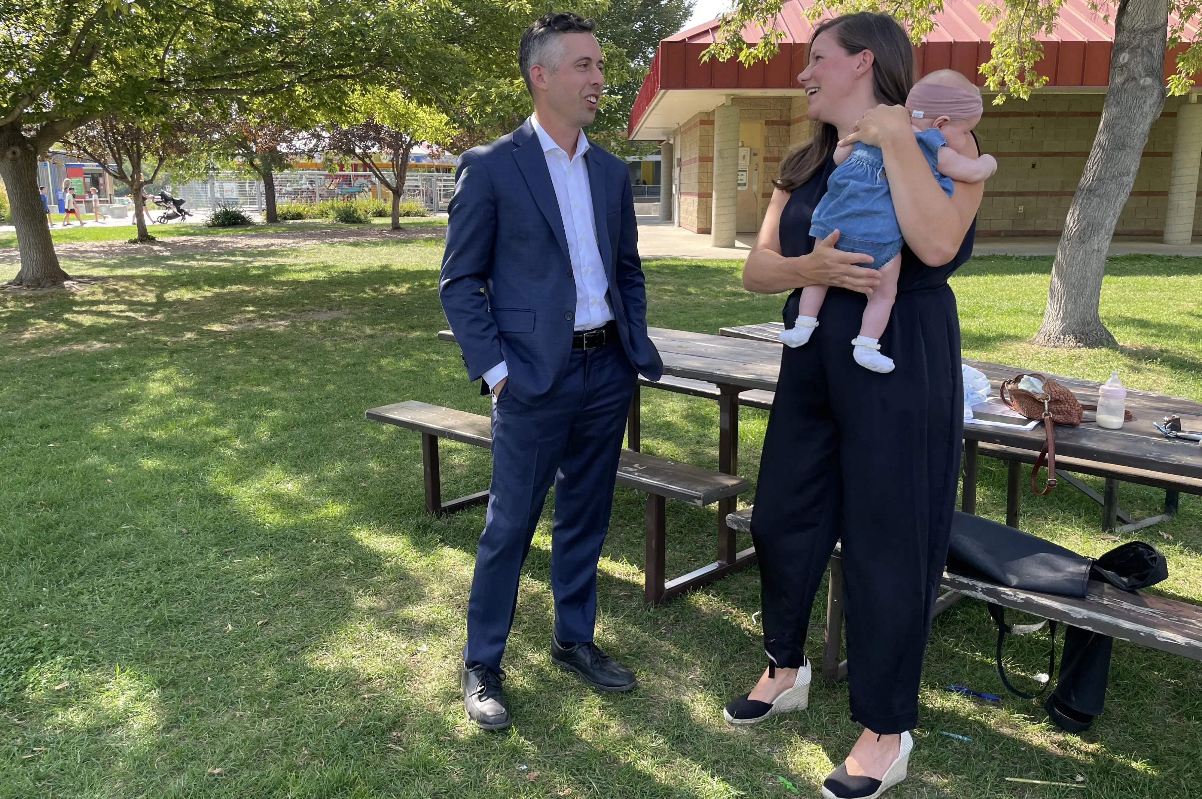 Jared Clarke, wearing a navy blue suit and light button-up shirt, talks with Meara Conway, wearing a black jumpsuit while holding her baby