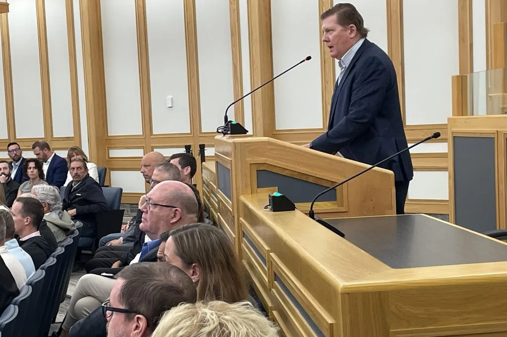 Keith Moen stands behind a podium looking, delivering his speech to city council while dozens of people fill the seats in front of him