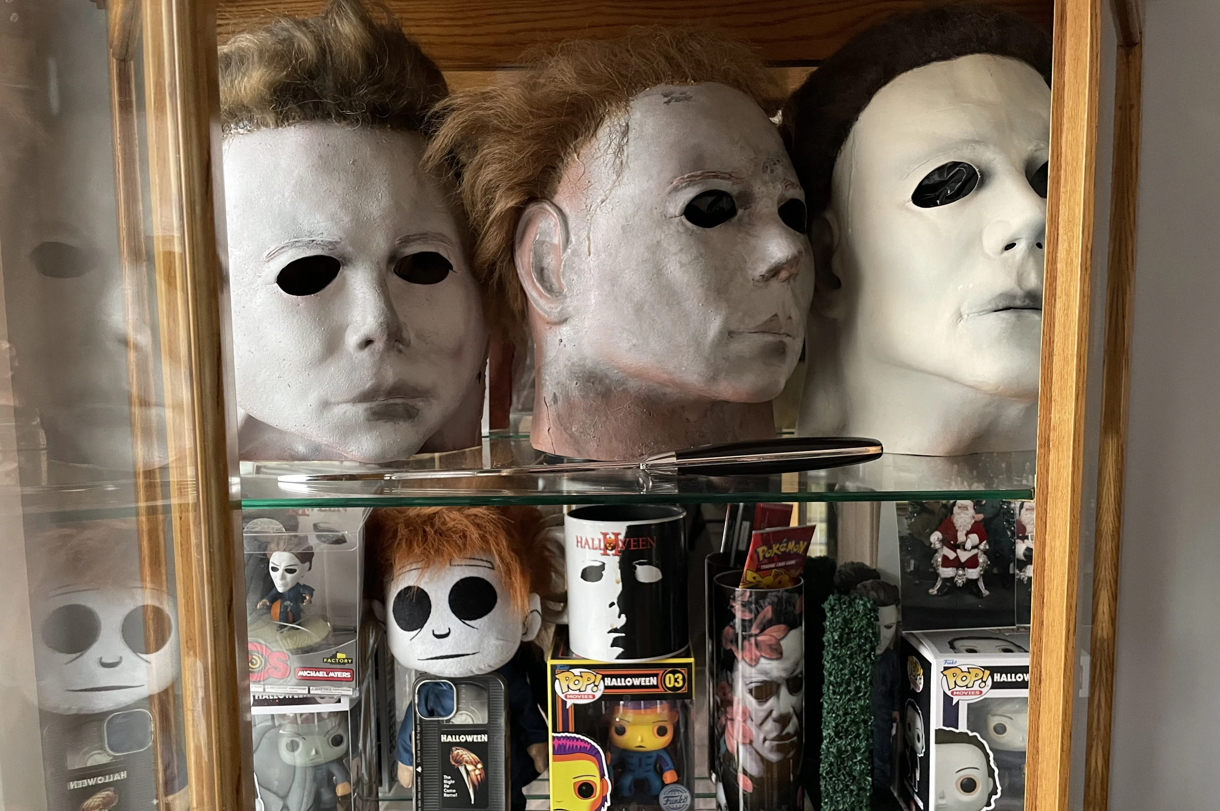 Three Michael Myers masks are on the top shelf in a display cabinet. They have white faces, cut outs for the eyes, and brown hair. In front of them is a knife. On the shelf below are various Myers collectible figurines still in their box.