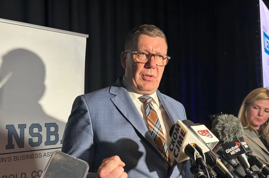 Premier Scott Moe speaks to reporters at Prairieland Park. A blond lady is in the back, to Moe's right. Several microphones are in front of the Premier, who stands in front of an NSBA banner. A reporter holds a phone in the foreground to record the scrum.
