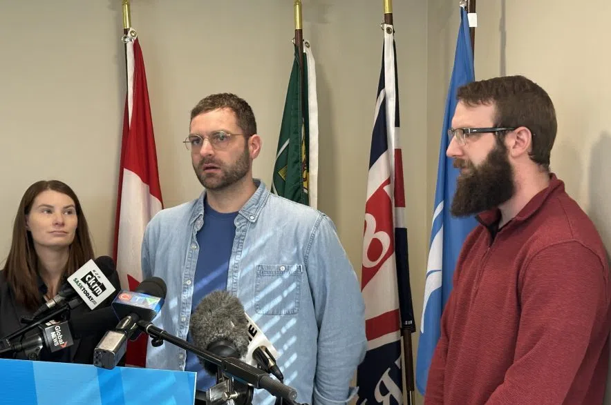 Alex Loewen and Timothy Carlow stand in front of flags and speak into microphones at the Saskatchewan NDP Saskatoon Riversdale office in Saskatoon. Kim Breckner, trade and export development critic, stands on the left.