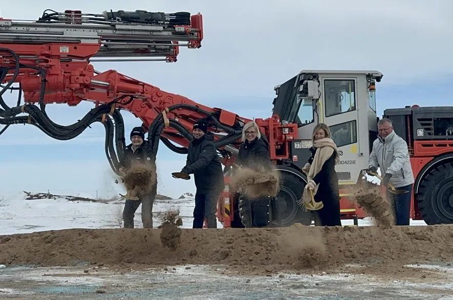 Five people toss sand and dirt with shovels in front of large, orange piece of machinery.