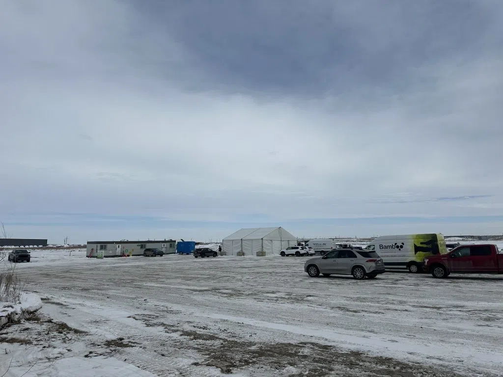 A white tent sits in the middle of the frame with parked cars nearby and a construction trailer. 