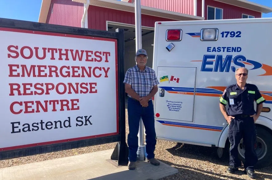 Donald, in a plaid t-shirt, jeans, glasses and a ball cap, stands by a sign on the left that says Southwest Emergency Response Centre, Eastend SK. To the right is the Eastend EMS ambulance and EMR Lloyd Weis in his paramedic uniform.