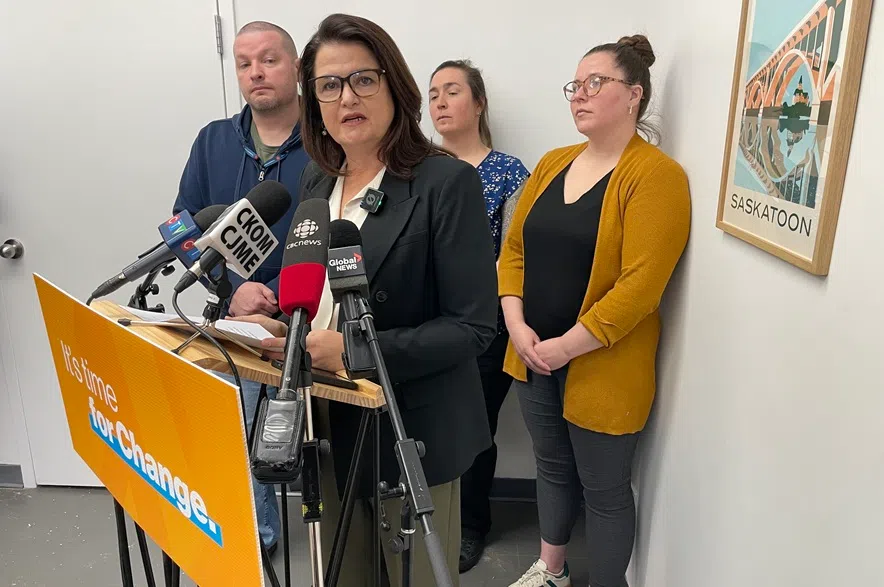 Carla Beck stands with Sarah Magnus, Matt Thompson and Christie MacNeill behind her. Beck is speaking into microphones on a podium with an orange NDP sign on it.