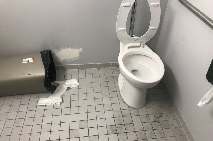 A toilet and tipped over garbage can in a dirty public bathroom.