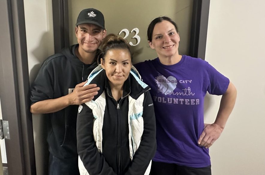 Tyler Rabbitskin (left) and Genine Okanee (middle) stand in front of their new apartment suite alongside Melissa Zimmer (right), founder of Bridge City Warmth, who helped the couple secure the housing. (Libby Gray/650 CKOM)