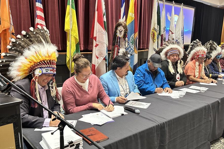 Saskatoon Tribal Council chief Mark Arcand (left), sits beside Minister of Indigenous Services Canada Mandy Gull-Masty and the seven chiefs of the tribal council's member nations on Oct. 7, 2026, as they all sign the new regional agreement concerning First Nations child welfare. (Libby Gray/650 CKOM)